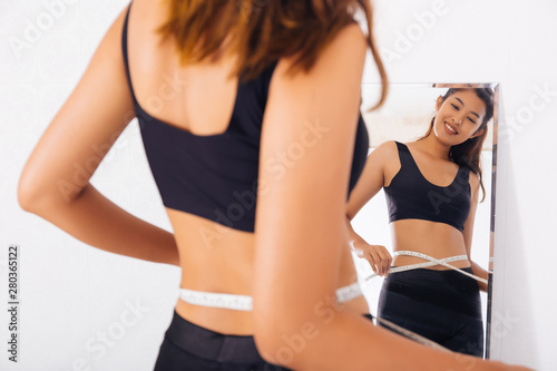 Young Asian woman measuring waist with tape in front of a mirror. Smiling sporty girl in sport bra looking at reflection. © twinsterphoto