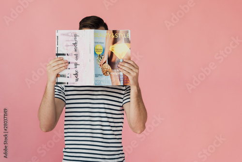 front view of man in striped t-shirt reading magazine isolated on pink