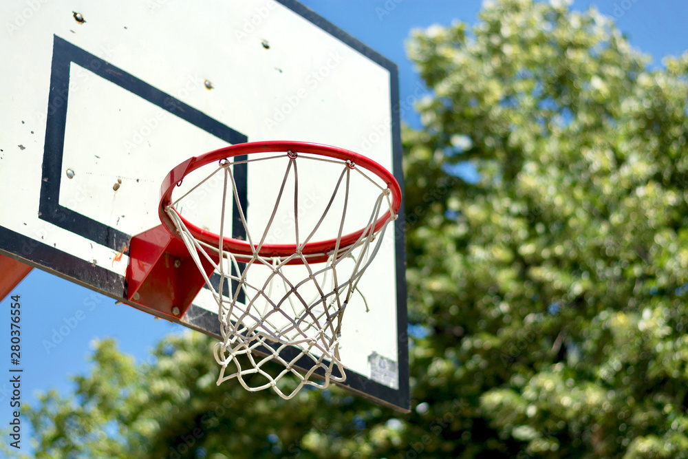 Side view of red metal basketball hoop with white net and backboard, outdoors, against the green park trees and cler blue sky background