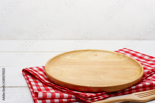 Empty round wooden plate with fork and red gingham tablecloth on white wooden table.