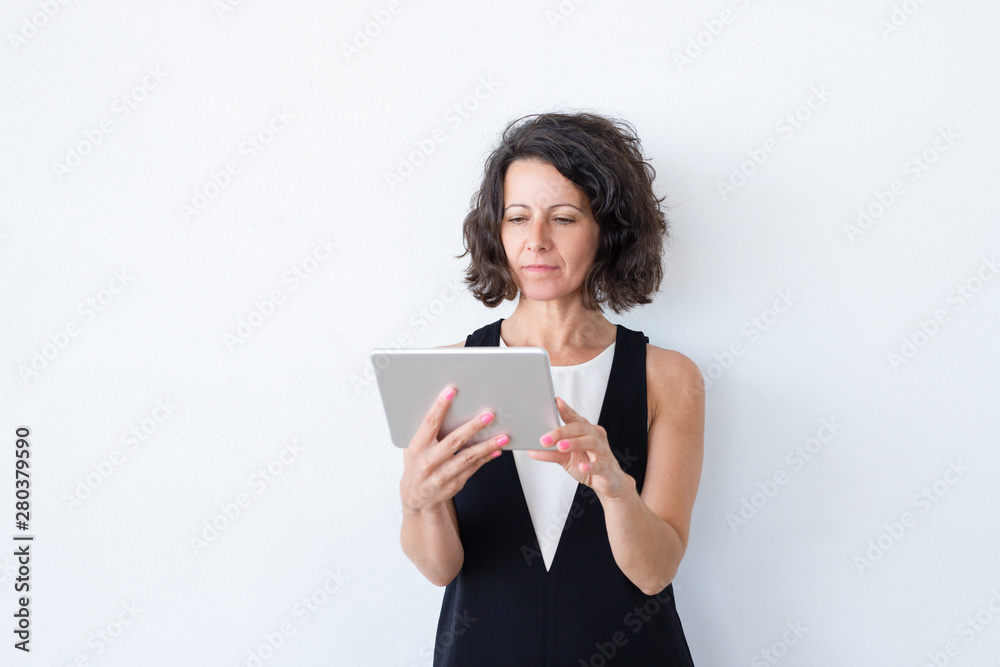 Serious focused woman in casual using tablet. Curly haired middle aged model standing over white background and reading on screen. Tablet using concept