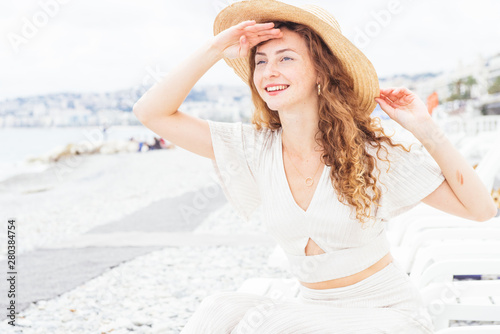 Beautiful ,elegant, teenager, with red hair and freckles,wearing a white dress and a hat, enjoy the seaside resort, watch in the distance the beautiful panorama covering her eyes with the palm of hand
