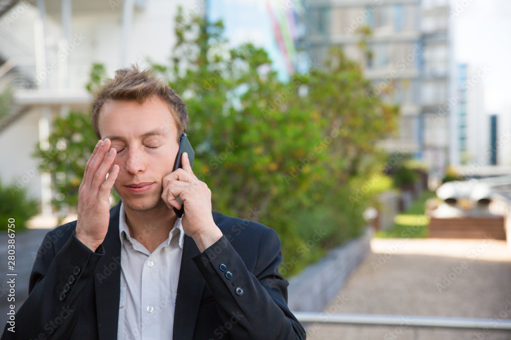 Tired business man calling on cellphone outside. Stressed office worker with closed eyes standing near city building, touching head and talking on cell. Stressful call concept Tired business man calling on cellphone outside. Stressed office worker with closed eyes standing near city building, touching head and talking on cell. Stressful call concept