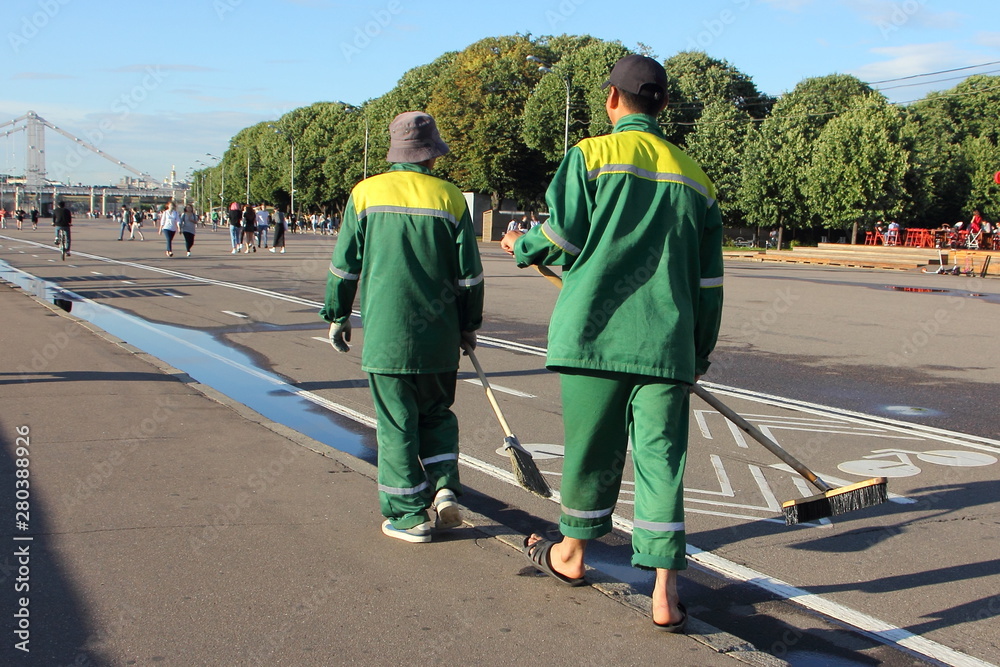 Two janitors with brooms and green yellow uniform are in Gorky Park on a Sunny summer day, municipal services of Moscow