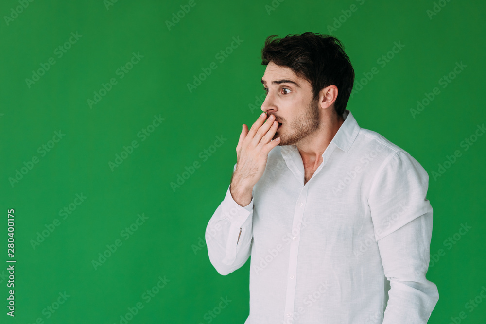 scared young man in white shirt covering mouth with hand isolated on green