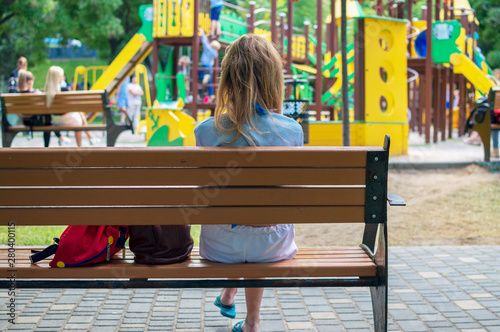 Young mother sits on a bench near the playground and looks after the child