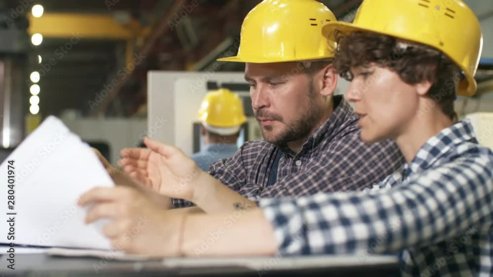 Pan chest-up shot of female and male factory engineers standing in modern automated production facility and studying data sheets with technical specifications, engaged in animated discussion Pan chest-up shot of female and male factory engineers standing in modern automated production facility and studying data sheets with technical specifications, engaged in animated discussion