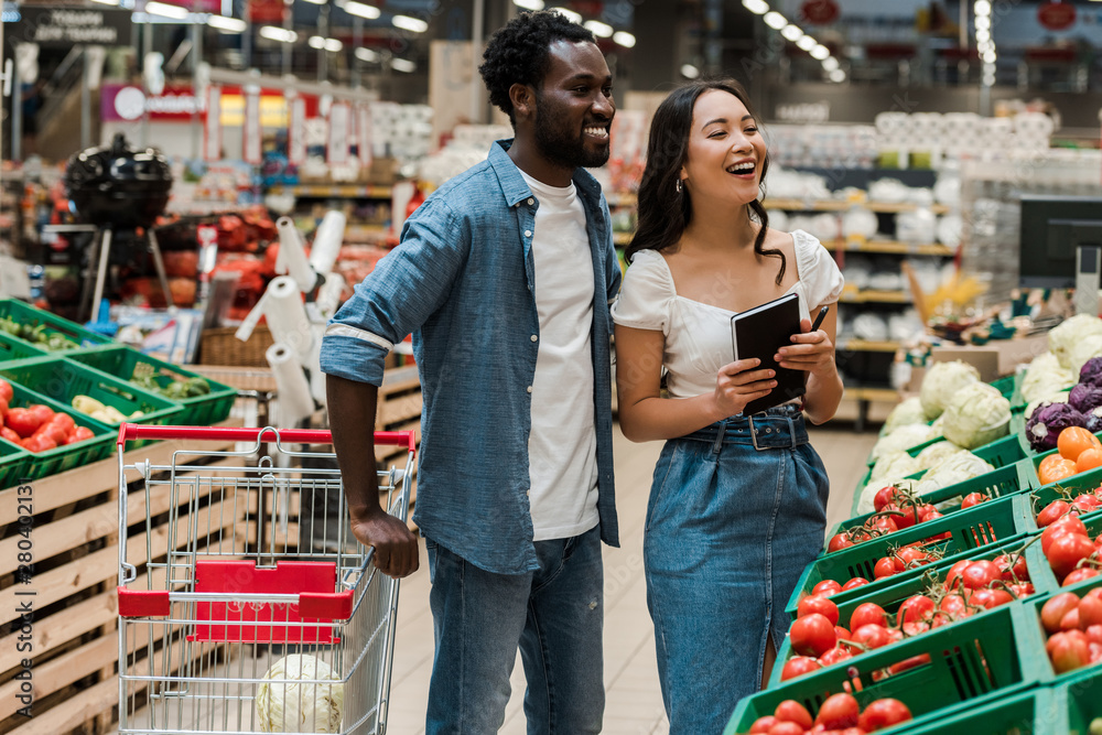 cheerful african american man near attractive asian woman holding notebook in supermarket