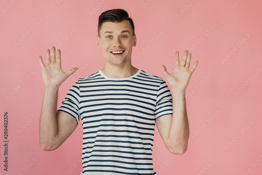 front view of excited smiling young man in striped t-shirt standing with hands up isolated on pink front view of excited smiling young man in striped t-shirt standing with hands up isolated on pink