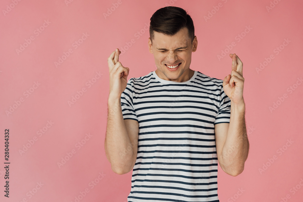 front view of worried young man in striped t-shirt with crossed fingers isolated on pink front view of worried young man in striped t-shirt with crossed fingers isolated on pink