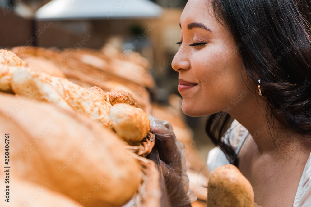 selective focus of cheerful asian woman smiling while smelling bread in supermarket - obrazy, fototapety, plakaty 