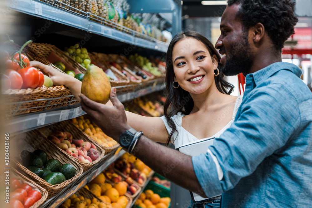 happy asian woman looking at african american man near fruits in store