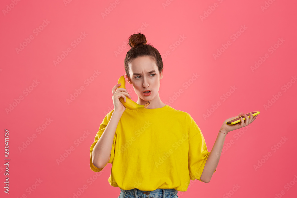 Confused model with a hair bun in a yellow t-shirt holding banana speaking on a yellow smartphone Confused model with a hair bun in a yellow t-shirt holding banana speaking on a yellow smartphone