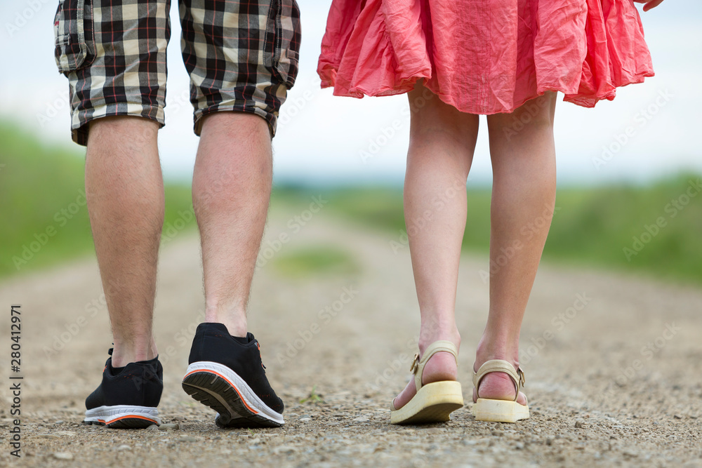 Back view of legs of young slim woman in red dress and man in shorts walking together by ground road on sunny summer day on blurred background. Back view of legs of young slim woman in red dress and man in shorts walking together by ground road on sunny summer day on blurred background.