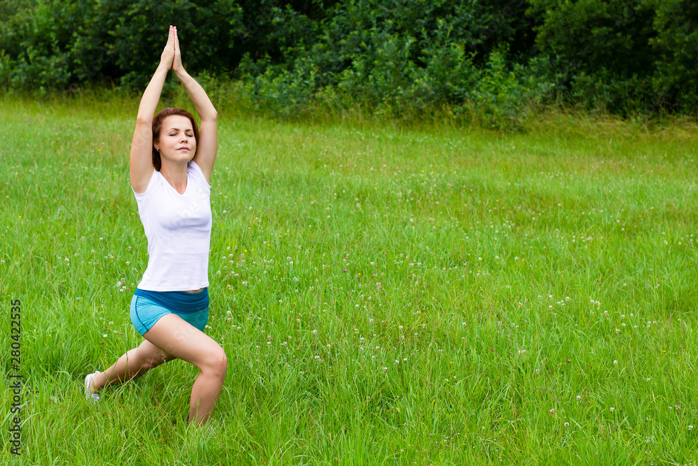 Woman practices yoga and meditates, girl doing pilates exercise standing in warrior one pose, virabhadrasana 1 in white T-shirt. Outside, in the forest, in the grass at a summer day. Front view. Woman practices yoga and meditates, girl doing pilates exercise standing in warrior one pose, virabhadrasana 1 in white T-shirt. Outside, in the forest, in the grass at a summer day. Front view.