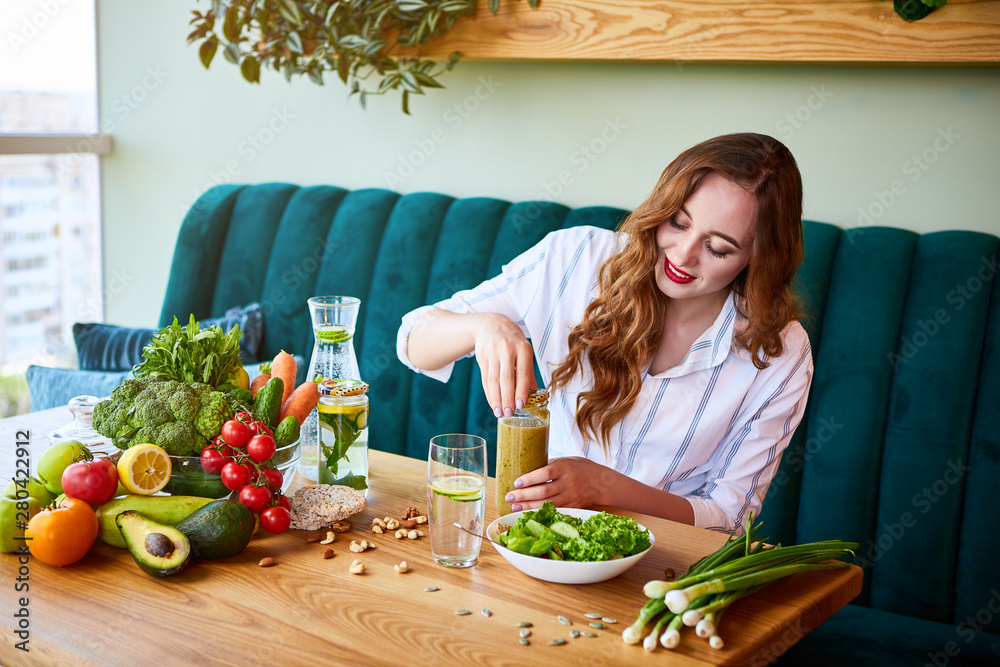 Young woman drinking smoothie in the beautiful interior with green flowers on the background and fresh fruits and vegetables on the table. Healthy eating concept. Vegan meal and detox menu