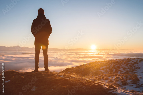 View From the back. A lonely standing man high in the mountains looks at the setting sun and the sunset horizon with a valley filled with clouds. The concept of tourism travel and male loneliness photo
