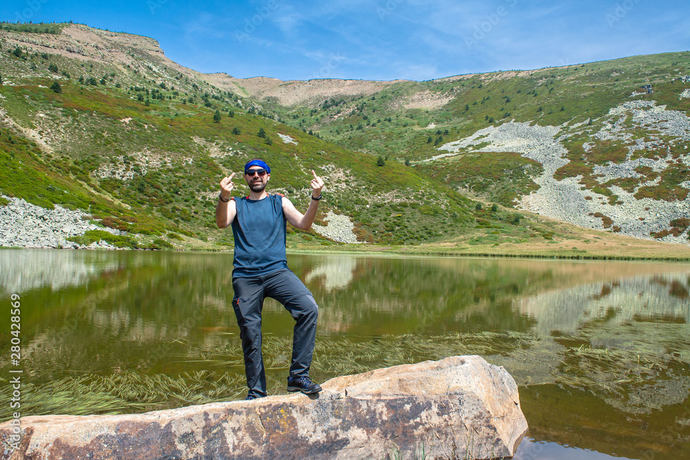 young angry man on rock in a mountain lake raises his arms
