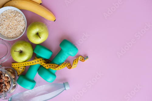 Fitness concept with dumbbells, nuts, bananas, apples and skipping rope on pink background with copy space