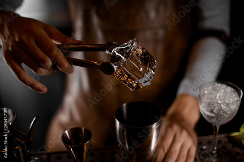 Bartender adds ice cube in cocktail with tongs