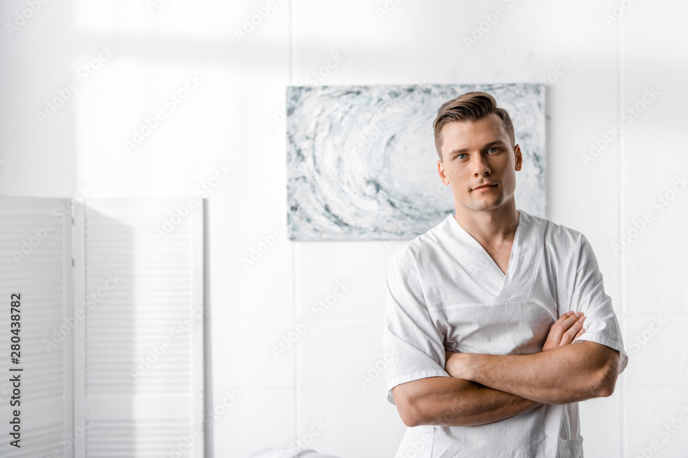 masseur standing with crossed arms and looking at camera in clinic
