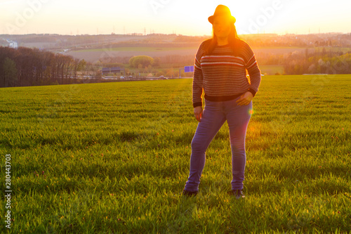 Lady poses outside standing at a filed at sunset for a portrait with the sun behind her. She is wearing a hat.