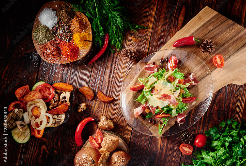 Salad with prosciutto, tomatoes, cheese, arugula and spinach in a transparent plate on dark wooden background in beautiful composition among vegetables and spices. Top view. Flat lay Salad with prosciutto, tomatoes, cheese, arugula and spinach in a transparent plate on dark wooden background in beautiful composition among vegetables and spices. Top view. Flat lay