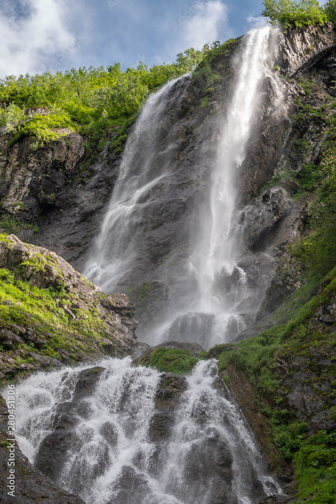 A high waterfall falls from a cliff in spring or summer.