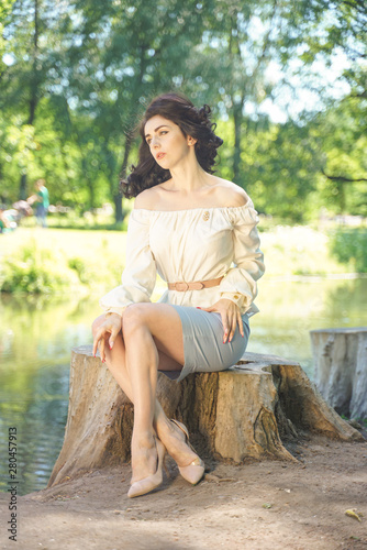 cute girl in a white summer dress tired of the sun and sat on a large stump near the lake in the shade of trees