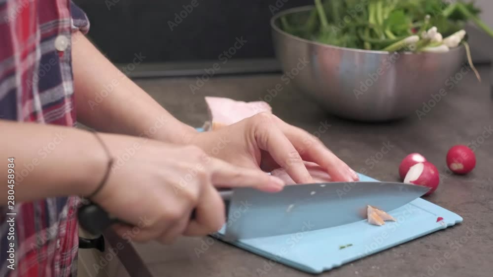 Young woman housewife cuts a piece of smoked pork ham on blue plastic cutting board. Preparing food ingredients on a stone desk at home kitchen. 4k 50 frames per second narrow depth of field close up.