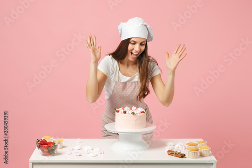 Housewife female chef cook confectioner or baker in apron white t-shirt, toque chefs hat cooking cake or cupcake at table isolated on pink pastel background in studio. Mock up copy space food concept.