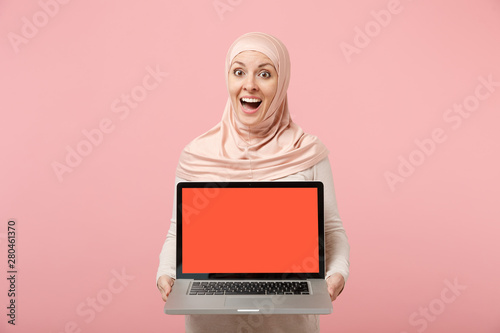 Surprised arabian muslim woman in hijab light clothes posing isolated on pink background. People religious Islam lifestyle concept. Mock up copy space. Hold laptop pc computer with blank empty screen.