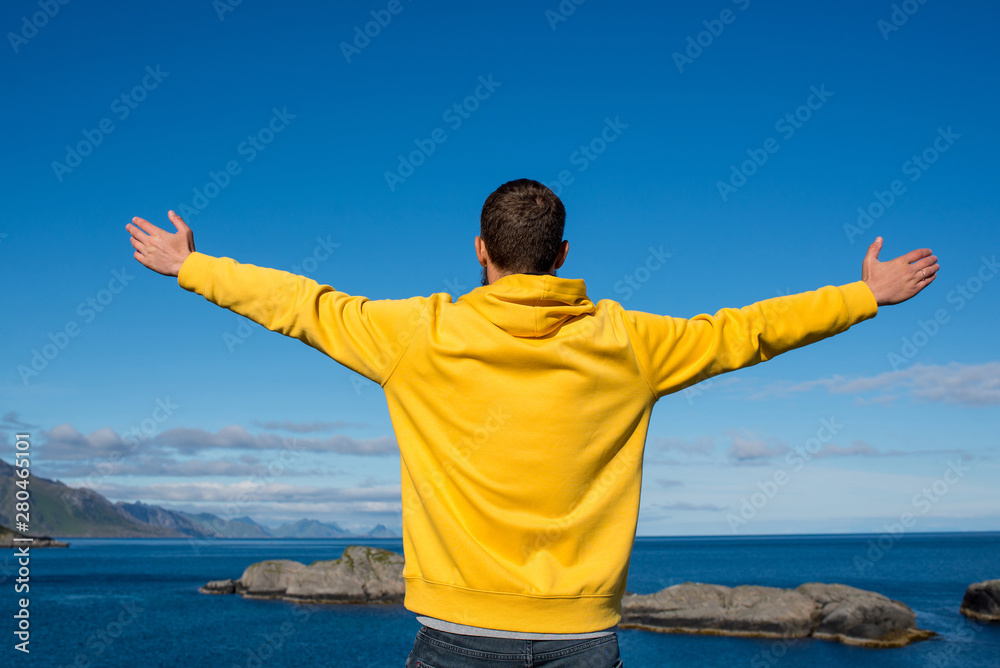 A man in a yellow hoodie outdoor with his arms raised against. Ocean and rocks landscape. Scenic view. Travel, adventure. Sense of freedom, lifestyle. Lofoten Islands, Norway. Summer in Scandinavia