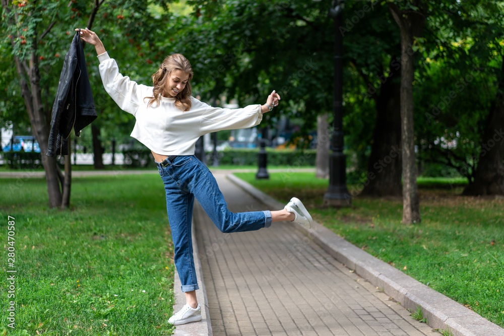Obraz premium beautiful young European girl walks in city green park. Caucasian girl posing standing on one leg. Lifestyle, pullover, jeans, sneakers. interesting pose full length in fashionable look.