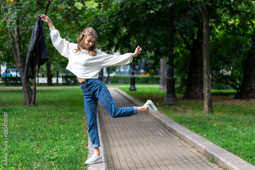 beautiful young European girl walks in city green park. Caucasian girl posing standing on one leg. Lifestyle, pullover, jeans, sneakers. interesting pose full length in fashionable look.