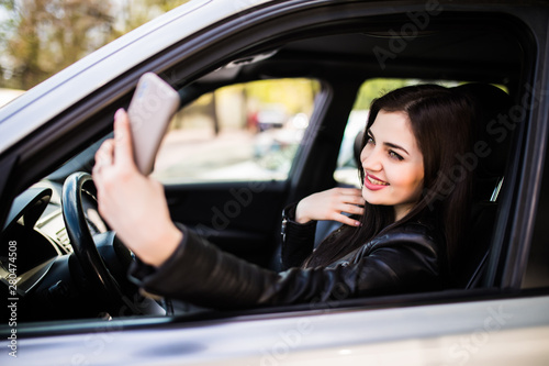 Happy young woman holding mobile phone and taking photos while driving a car. Smiling girl taking selfie picture with smart phone camera outdoors in car. Holidays and tourism concept © dianagrytsku