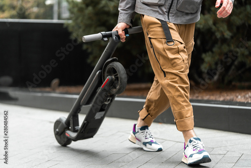 Close up of man holding electric kick scooter while going for a walk at street.