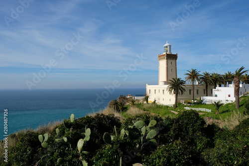 In Morocco Tangier white lighthouse near blue ocean. Sunny clouds skyline . Green cactus foreground. Cape Malabata with a lighthouse. promontory at the entrance to the Strait of Gibraltar