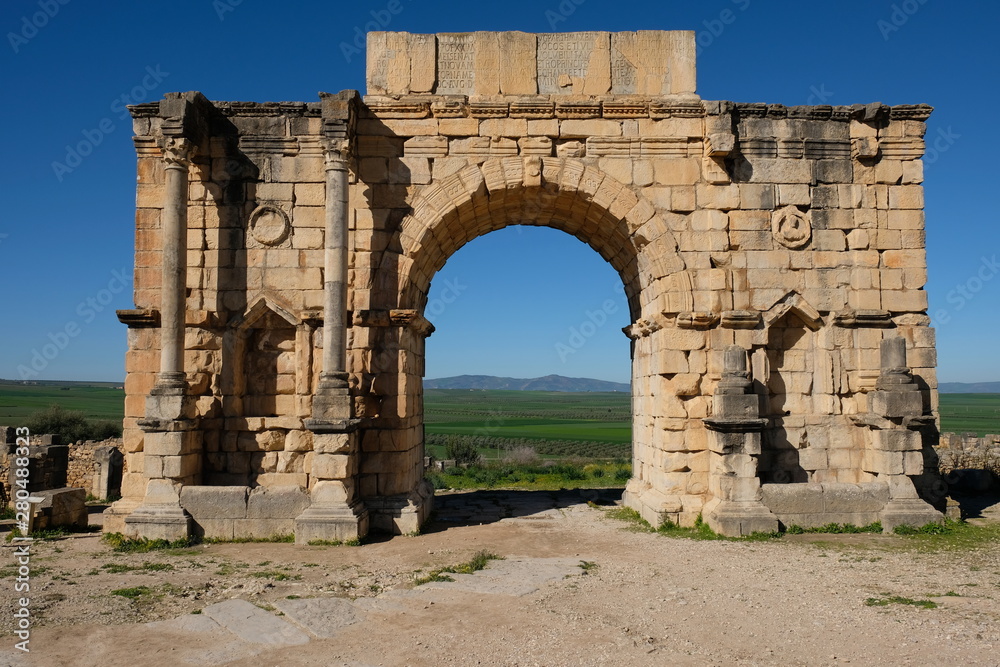 Roman Ruins brown arched relic gate in volubilis in morocoo.  The Triumphal Arch gate. Stone brick ruins of Roman capital. green grassland and blue sunny sky background