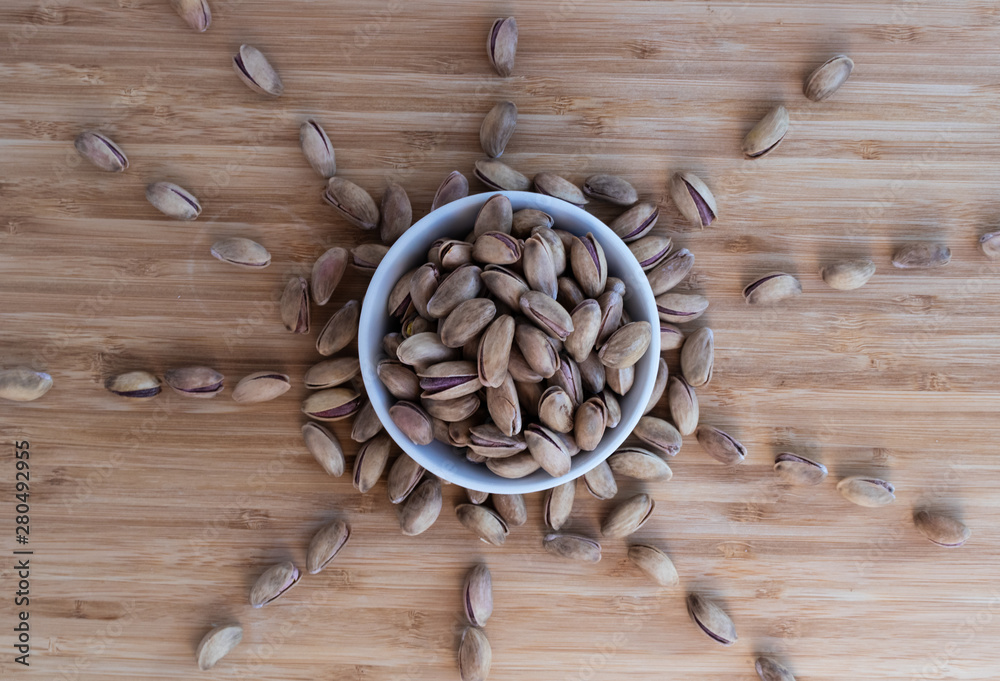 Pistachio nuts piled to the top in a white bowl on a light brown background, top view