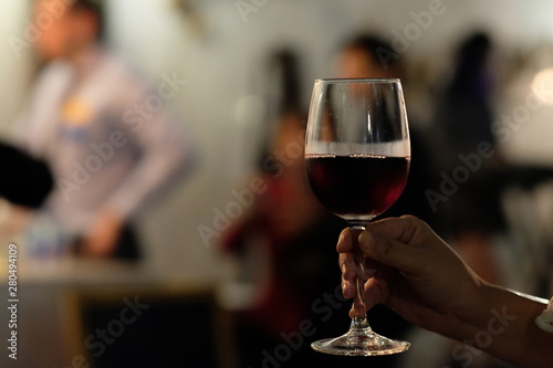 close up one hand holding a red wine glass with defocused people and light background