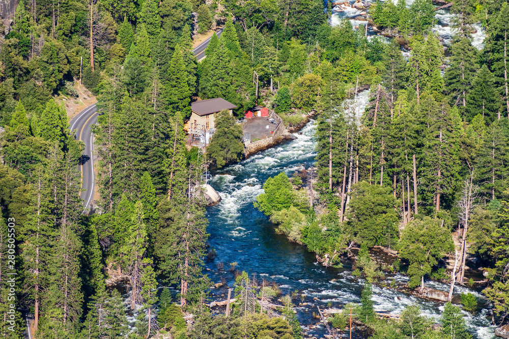 Aerial view of Merced river flowing through a coniferous forest; electric power station and paved road visible on the left; Yosemite National Park, California