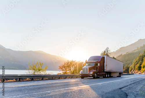 Big rig burgundy semi truck transporting commercial cargo in refrigerator semi trailer driving on the road along Columbia River with sunshine photo