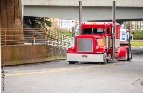 Classic stylish big rig red semi truck transporting cargo on flat bed semi trailer running under the bridge