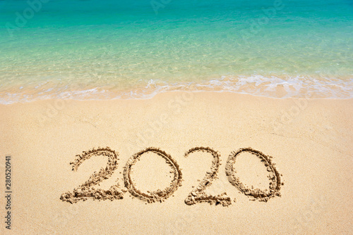 Happy New Year 2020, lettering on the beach with wave and clear blue sea. Numbers 2020 year on the sea shore, message handwritten in the golden sand on beautiful beach background. New Years concept.