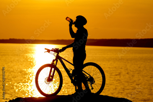 Silhouette of cyclist. Sporty guy cycling in helmet drinks water on the natural background of setting the sun in the evening.