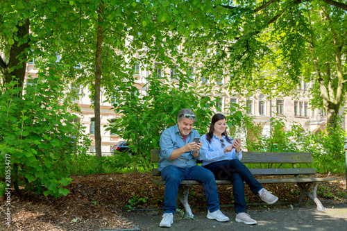 Couple sitting on bench of a city park. Each one is texting on the phone