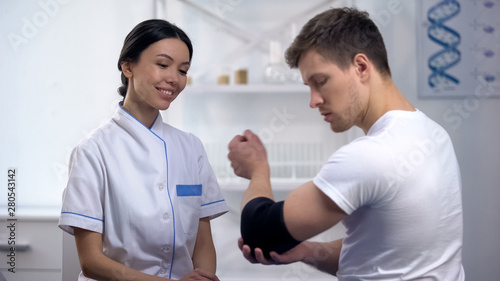 Male patient checking elbow padded orthosis, female orthopedist smiling, clinic
