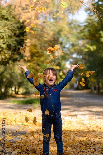 Beautiful little girl in denim shirt and jeans is sitting on yellow leaves and playing on warm and sunny autumn day. Happy childhood concept