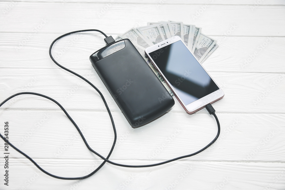 Black power bank with usb and smartphone on wooden background. Dollar notes lying nearby. Modern information technology photo.